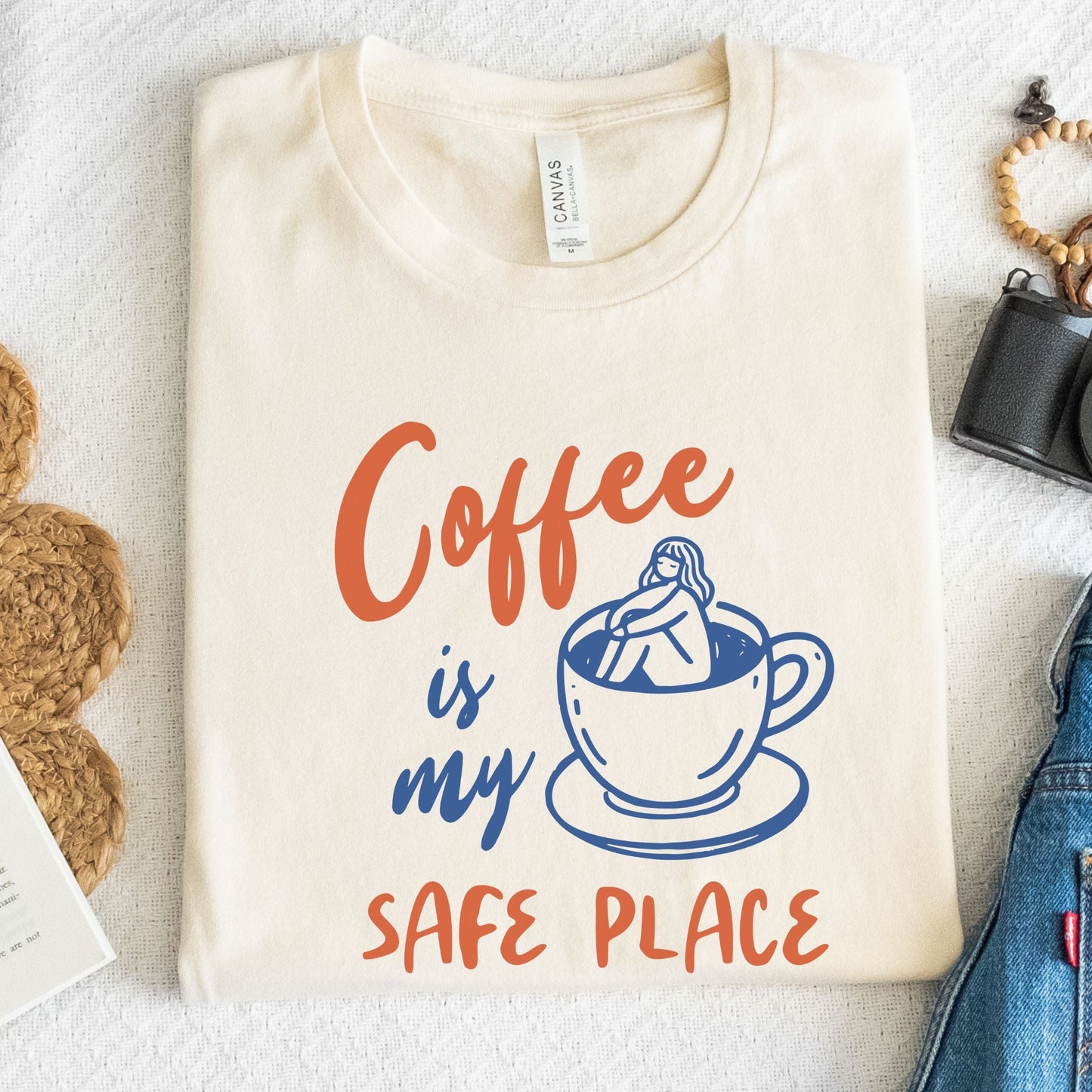 Blue and orange graphic t-shirt with text “Coffee is my safe place” featuring a girl sitting inside a coffee cup illustration.