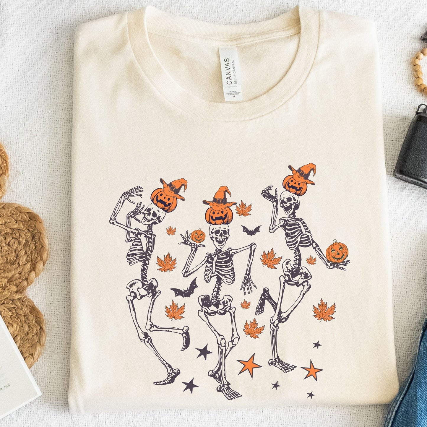 Halloween t-shirt with three dancing skeletons wearing pumpkin heads and witch hats, surrounded by autumn leaves, bats, and stars.