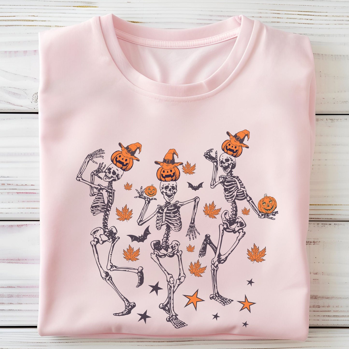 Halloween t-shirt with three dancing skeletons wearing pumpkin heads and witch hats, surrounded by autumn leaves, bats, and stars.