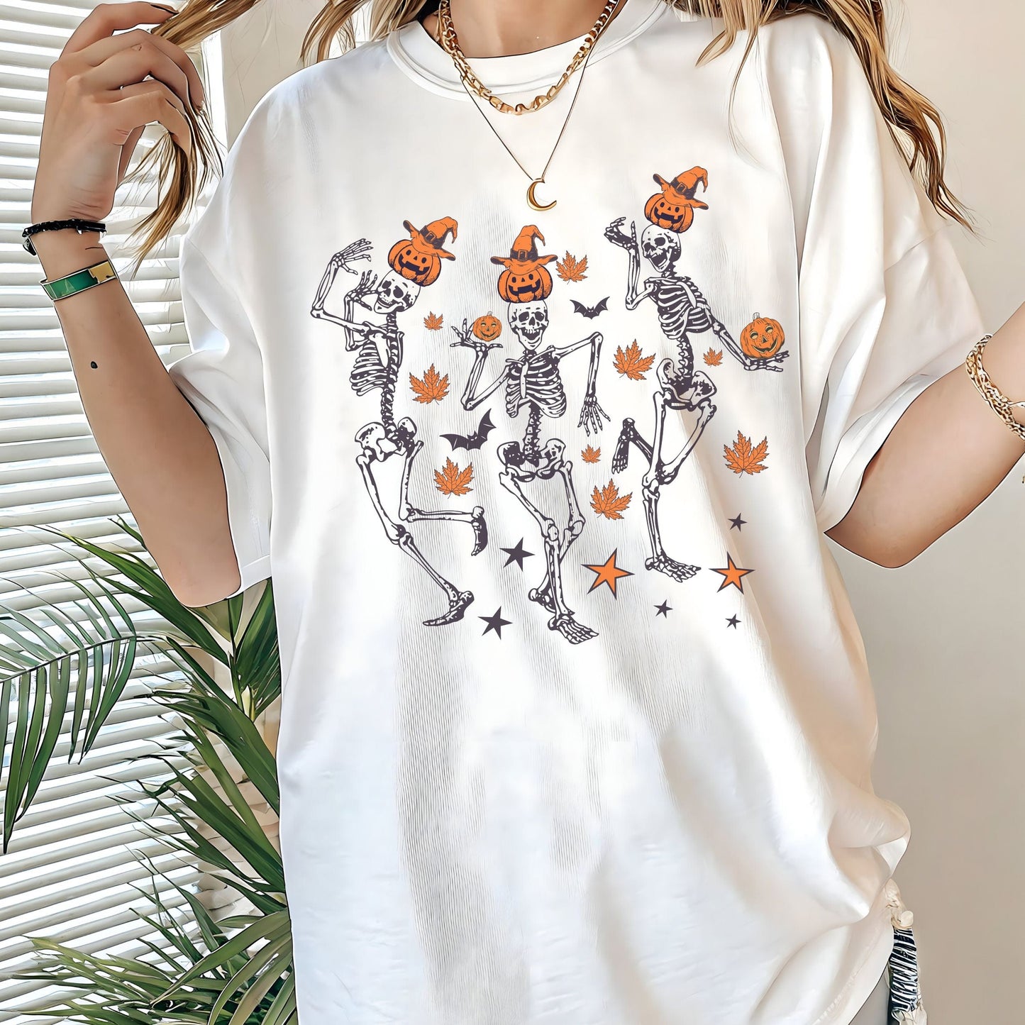 Halloween t-shirt with three dancing skeletons wearing pumpkin heads and witch hats, surrounded by autumn leaves, bats, and stars.