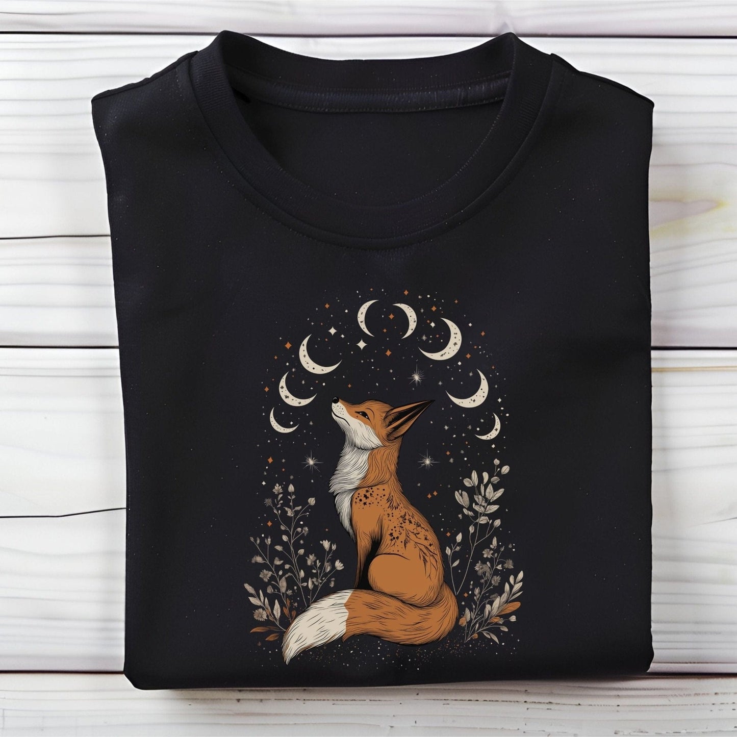 Celestial fox t-shirt with a woodland fox sitting under moon phases, surrounded by stars and hand-drawn floral illustrations in neutral tones.