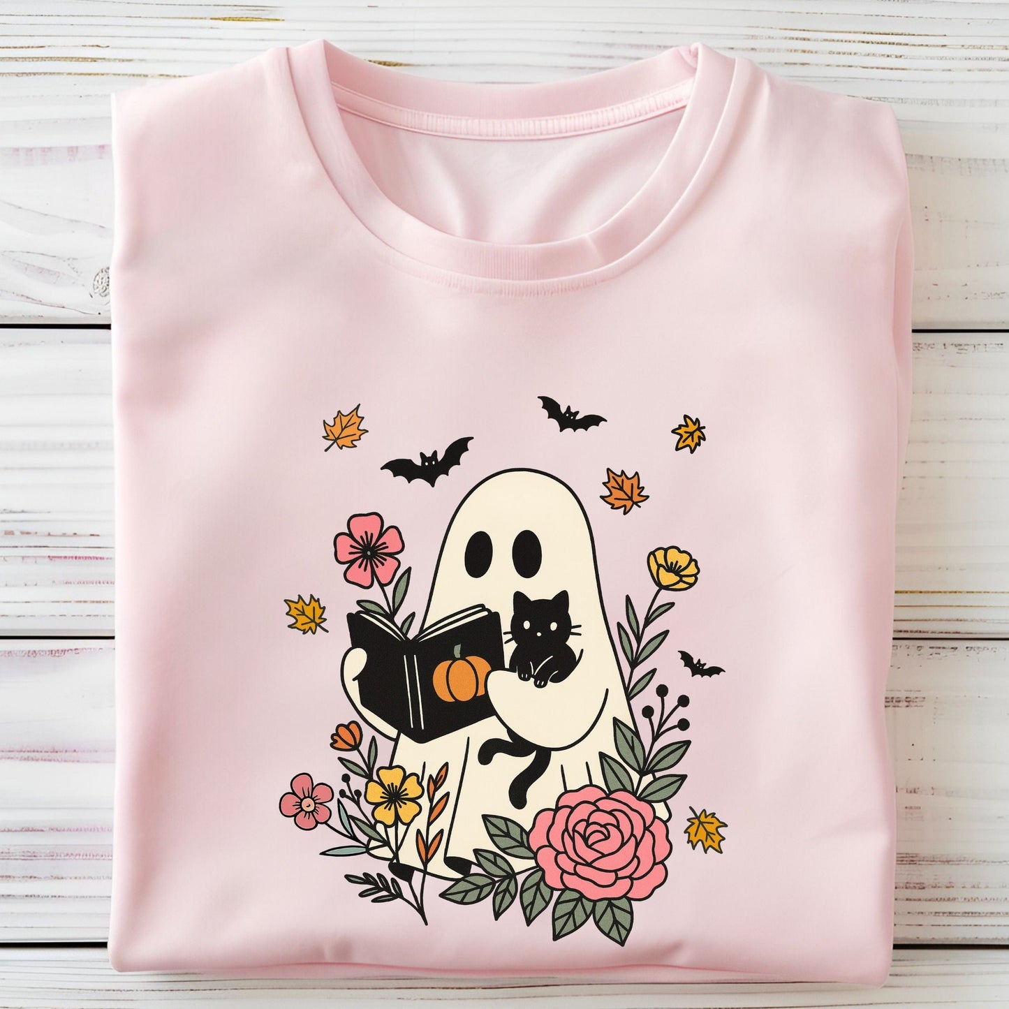 Folded pink t-shirt with Halloween-themed design featuring a ghost, cat, and pumpkins on a wooden surface.