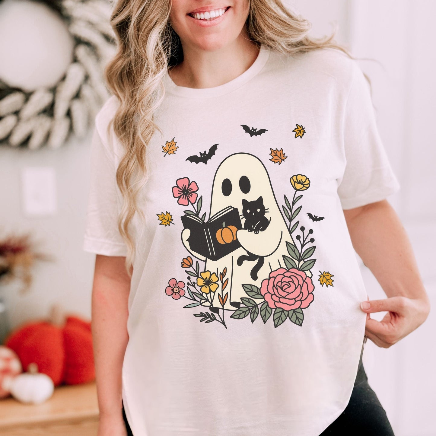 Person wearing a white t-shirt with a Halloween-themed design featuring a ghost, black cat, and flowers.