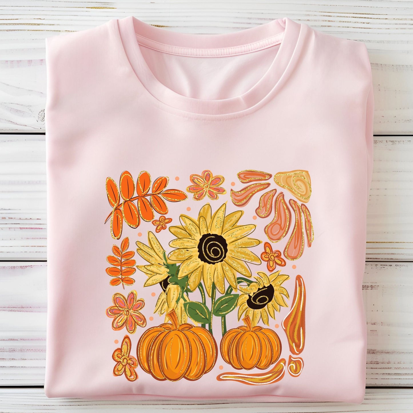 Folded soft pink t-shirt with autumn-themed design featuring sunflowers and pumpkins on a wooden surface.