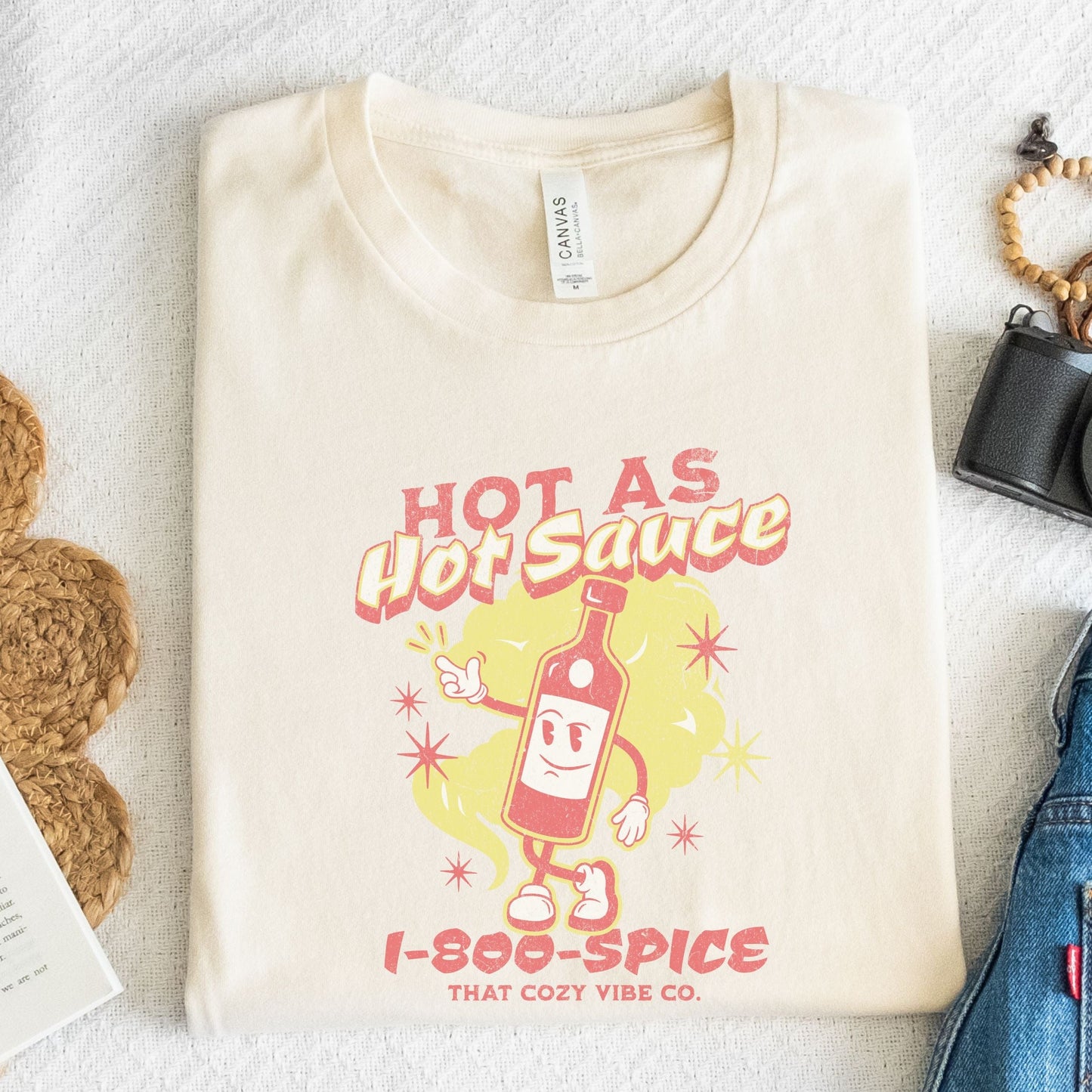 “Hot As Hot Sauce” t-shirt with retro cartoon hot sauce bottle character, bold yellow and red text, spice cloud, and “1-800-SPICE” at the bottom.