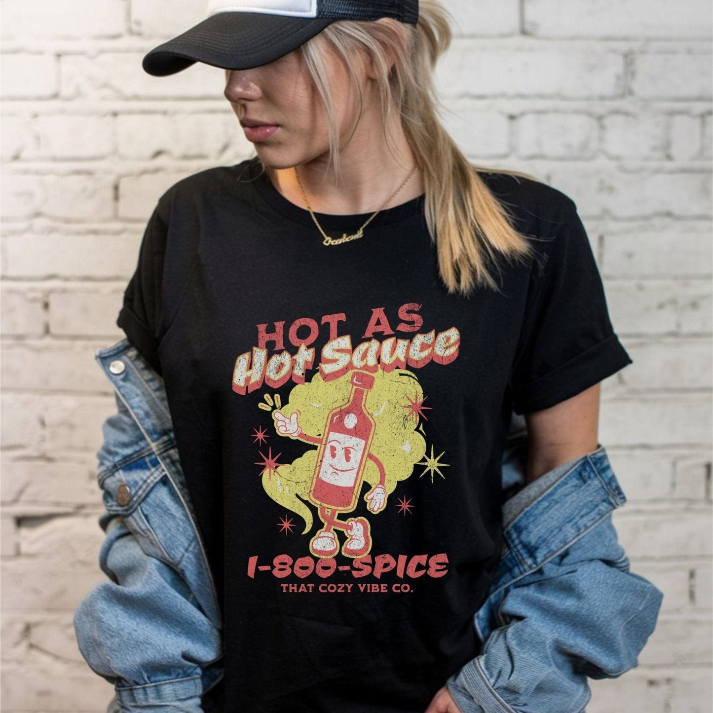 “Hot As Hot Sauce” t-shirt with retro cartoon hot sauce bottle character, bold yellow and red text, spice cloud, and “1-800-SPICE” at the bottom.