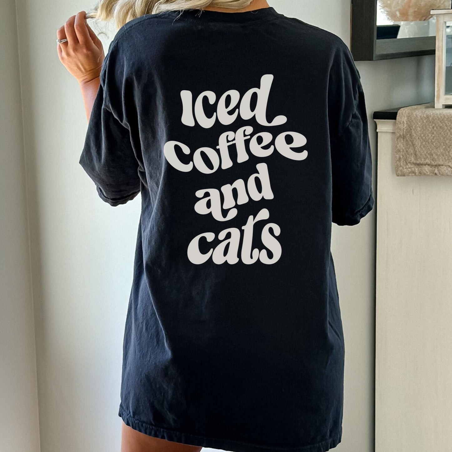 T-shirt with “Iced Coffee and Cats” in bold lettering, modeled in a cozy indoor setting.