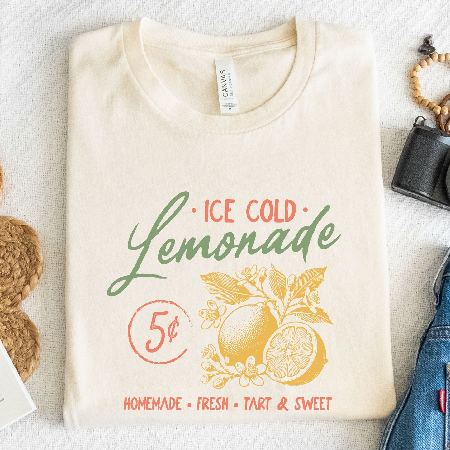T-shirt design featuring vintage-style "Ice Cold Lemonade" text with yellow lemons, floral details, and a 5¢ sign for a retro summer vibe.