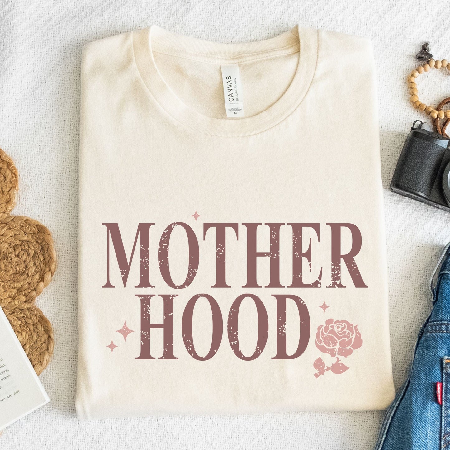 T-shirt with “Motherhood” text in distressed font and floral graphic. A soft, meaningful gift tee for moms.