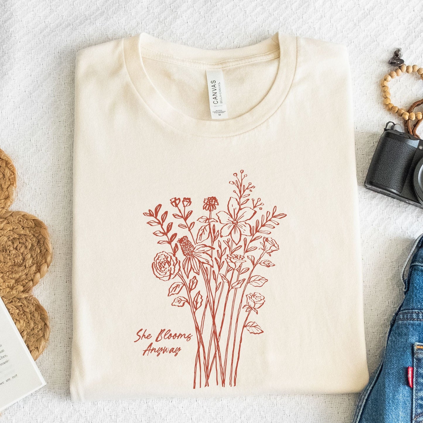 Floral graphic t-shirt with hand-drawn wildflowers and the quote “She Blooms Anyway”