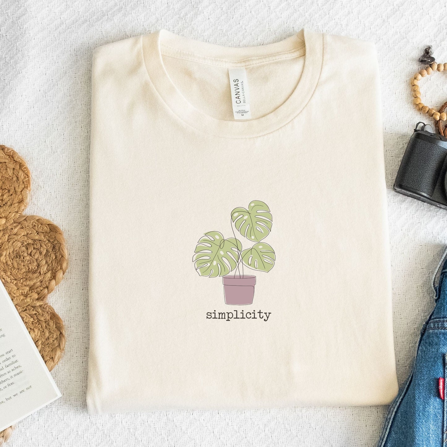 “Simplicity” t-shirt with minimalist monstera plant in a muted pink pot and soft, modern lettering beneath the design.