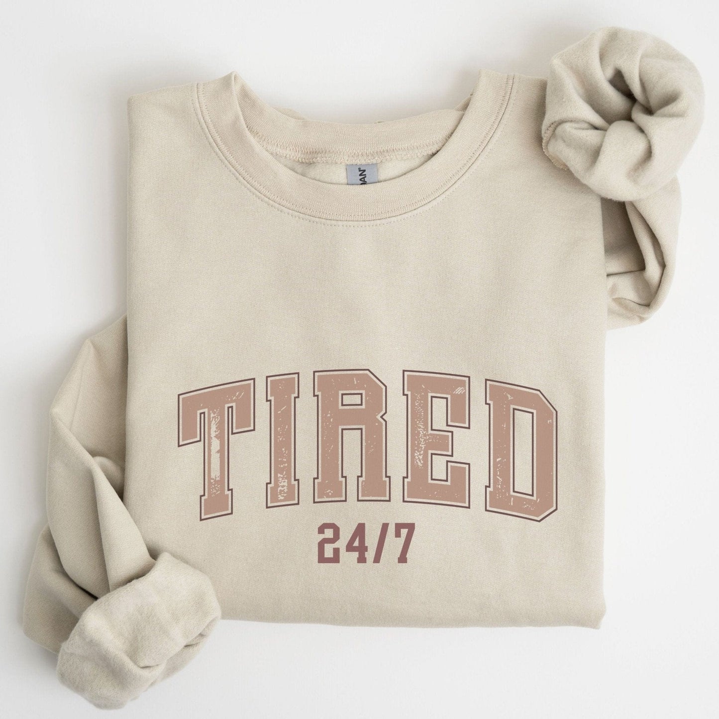 “Tired 24/7” sweatshirt with varsity block letters and a distressed texture — cozy and relatable for the chronically exhausted.
