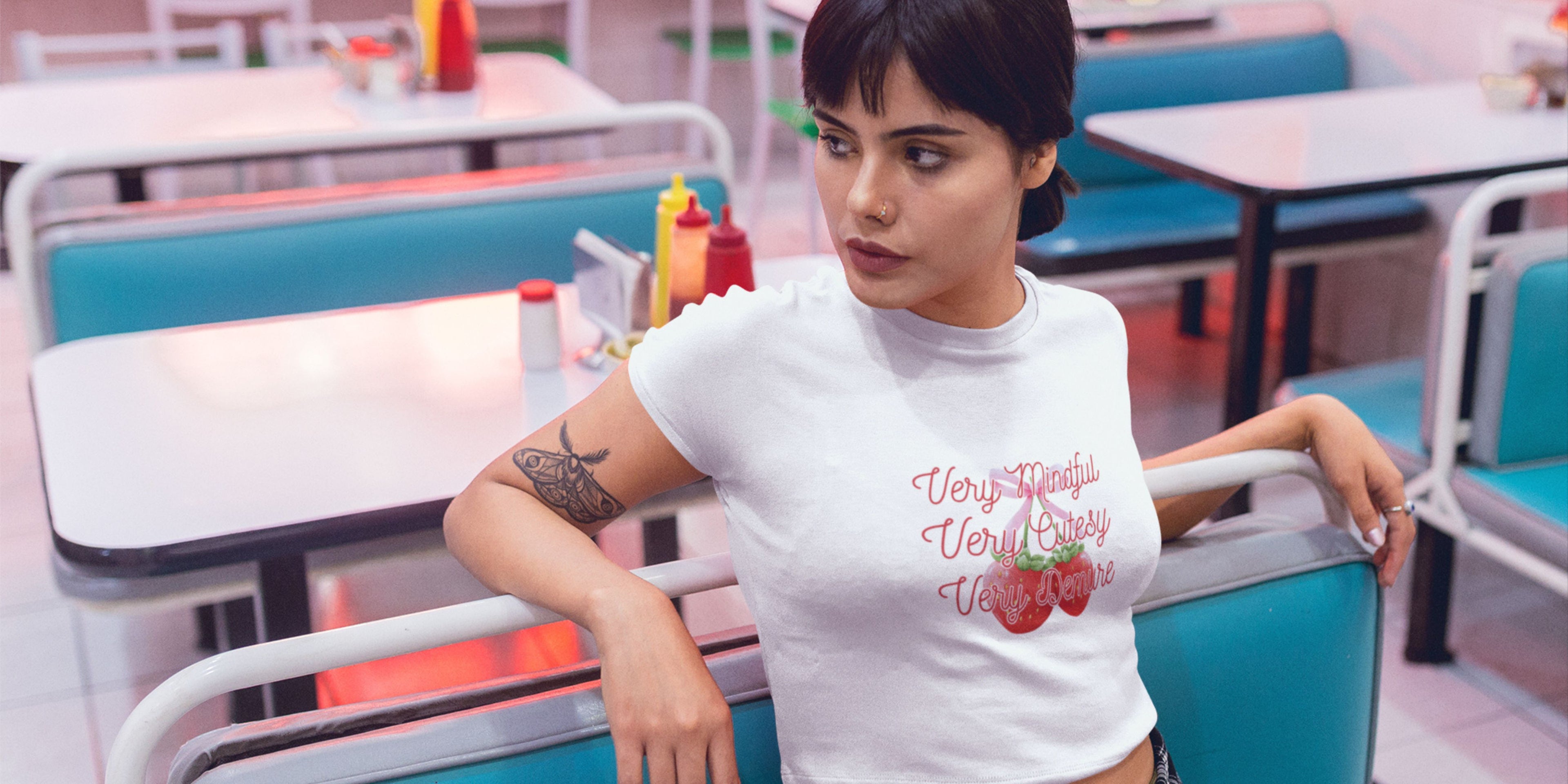 Model wearing a white baby tee with 'Very Mindful, Very Cutesy, Very Demure' text in a retro diner.