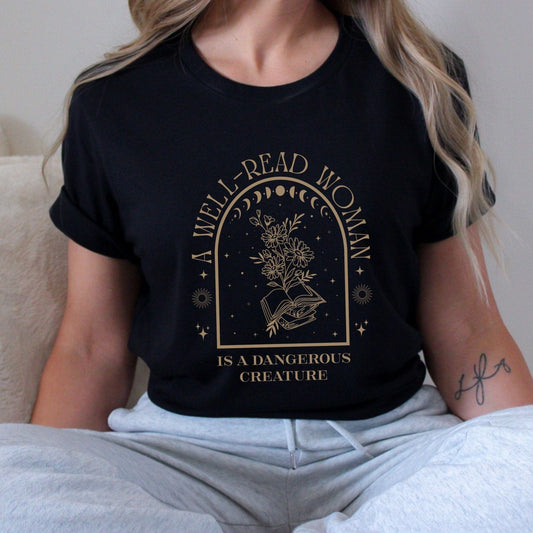“A Well-Read Woman Is a Dangerous Creature” t-shirt featuring celestial elements, moon phases, blooming wildflowers, and open books in a mystical arch design.