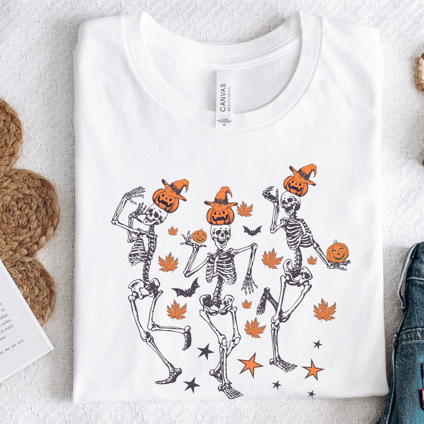 Halloween t-shirt with three dancing skeletons wearing pumpkin heads and witch hats, surrounded by autumn leaves, bats, and stars.
