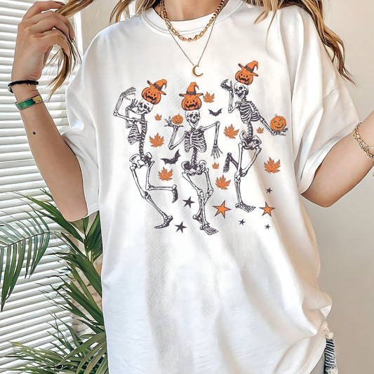Halloween t-shirt with three dancing skeletons wearing pumpkin heads and witch hats, surrounded by autumn leaves, bats, and stars.