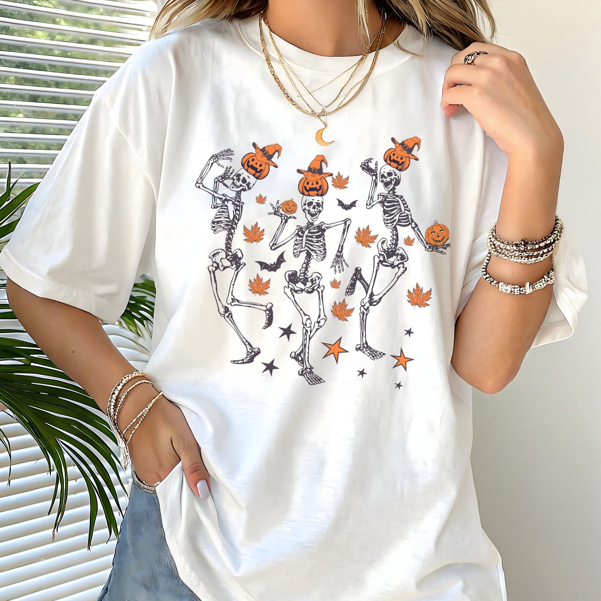 Halloween t-shirt with three dancing skeletons wearing pumpkin heads and witch hats, surrounded by autumn leaves, bats, and stars.