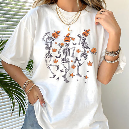Halloween t-shirt with three dancing skeletons wearing pumpkin heads and witch hats, surrounded by autumn leaves, bats, and stars.
