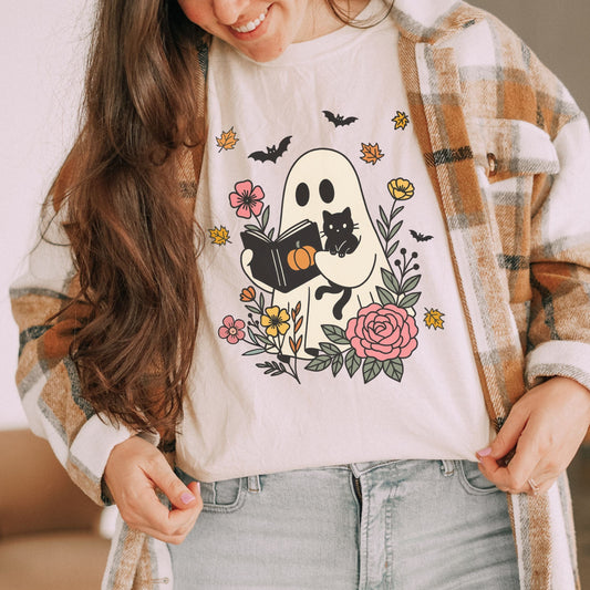 Person wearing a white t-shirt with a ghost holding a cat and reading with florals design, plaid jacket, and jeans.