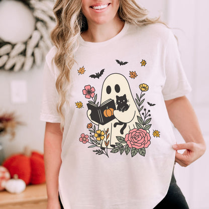 Person wearing a white t-shirt with a Halloween-themed design featuring a ghost, black cat, and flowers.