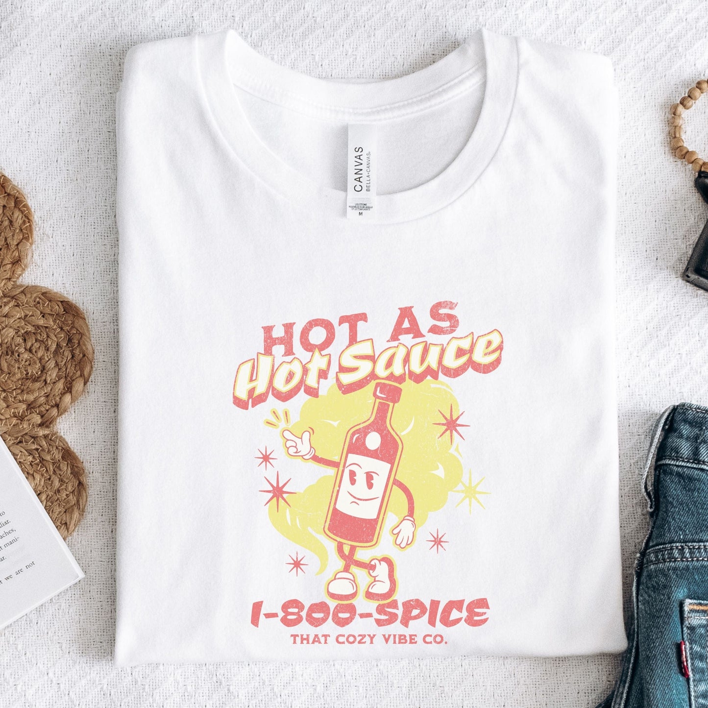“Hot As Hot Sauce” t-shirt with retro cartoon hot sauce bottle character, bold yellow and red text, spice cloud, and “1-800-SPICE” at the bottom.