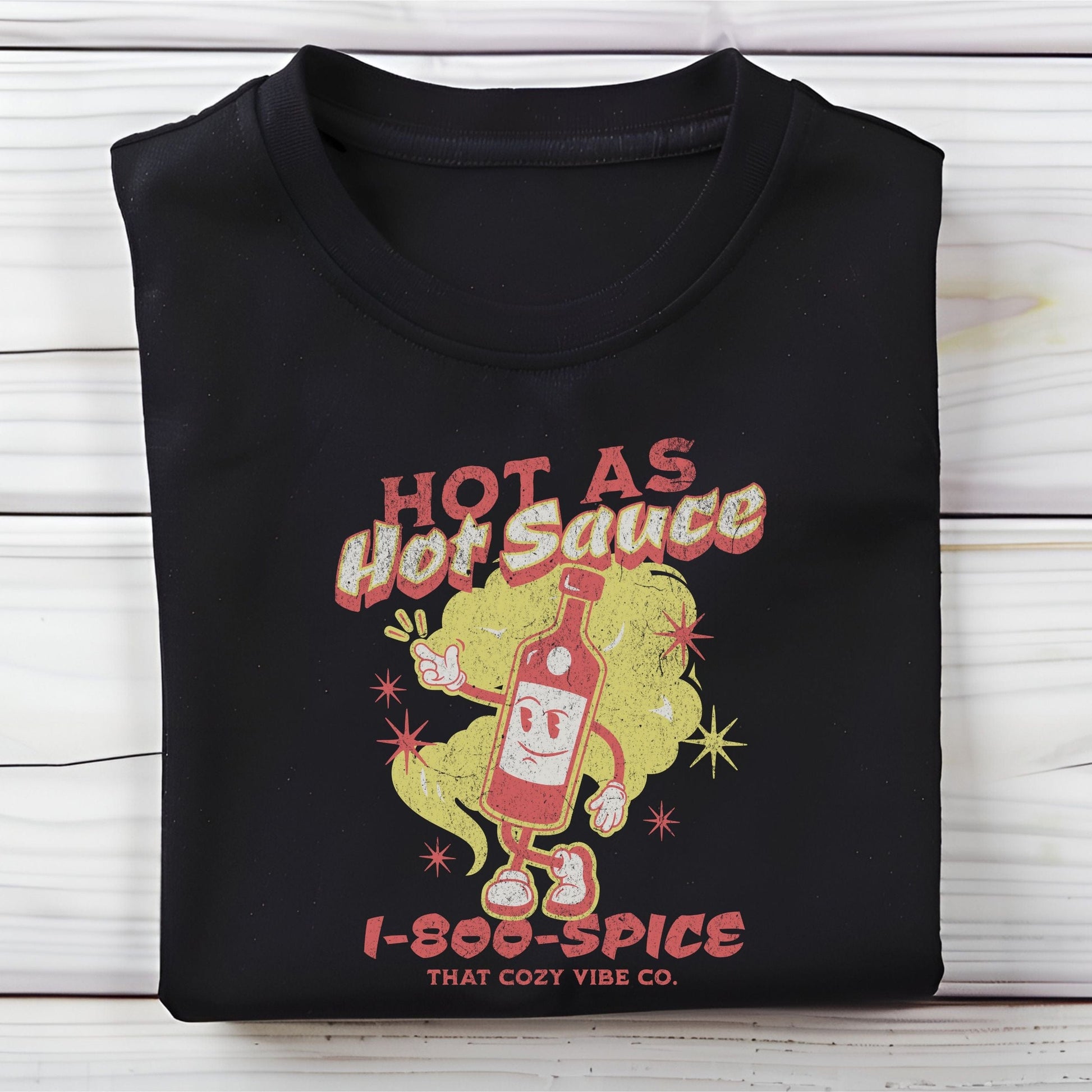“Hot As Hot Sauce” t-shirt with retro cartoon hot sauce bottle character, bold yellow and red text, spice cloud, and “1-800-SPICE” at the bottom.