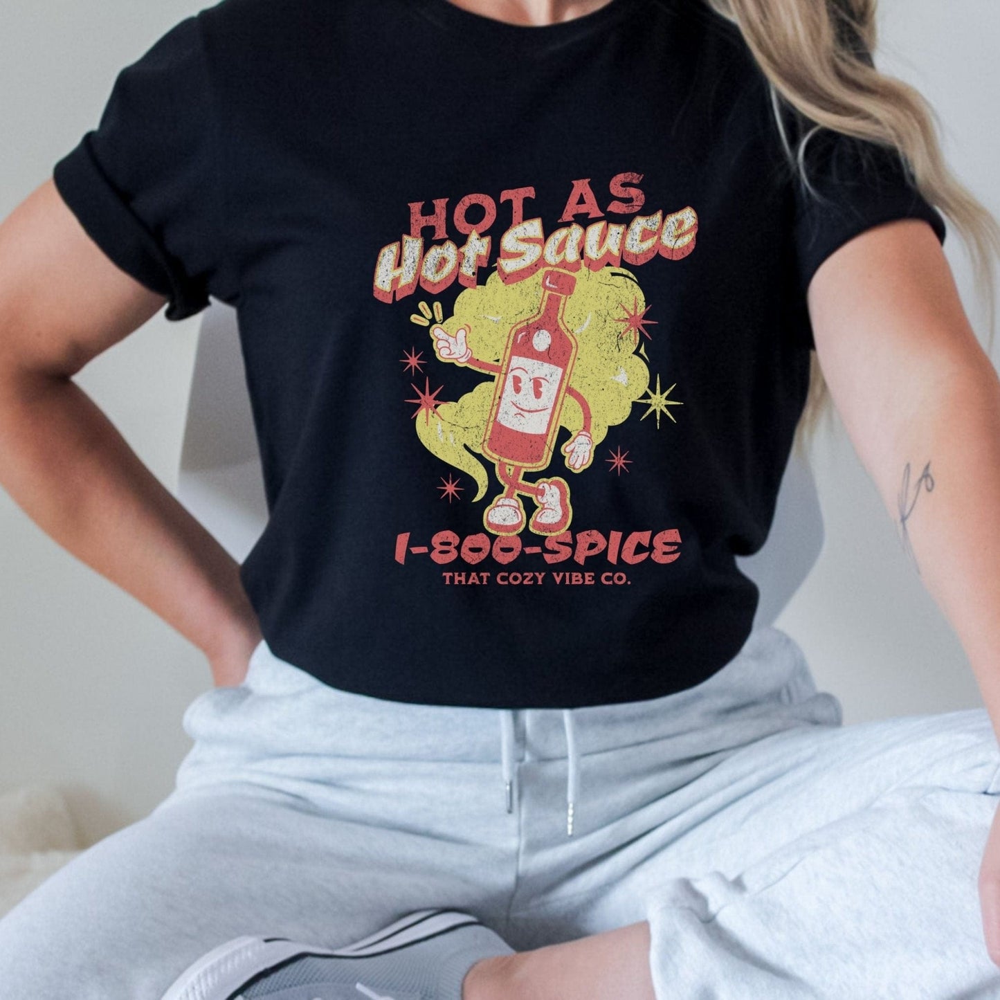 “Hot As Hot Sauce” t-shirt with retro cartoon hot sauce bottle character, bold yellow and red text, spice cloud, and “1-800-SPICE” at the bottom.