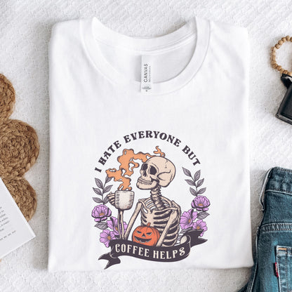“I Hate Everyone But Coffee Helps” t-shirt with skeleton drinking coffee, purple flowers, jack-o’-lantern, and gothic Halloween design.