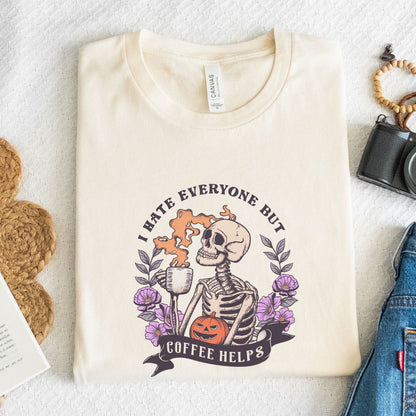 “I Hate Everyone But Coffee Helps” t-shirt with skeleton drinking coffee, purple flowers, jack-o’-lantern, and gothic Halloween design.