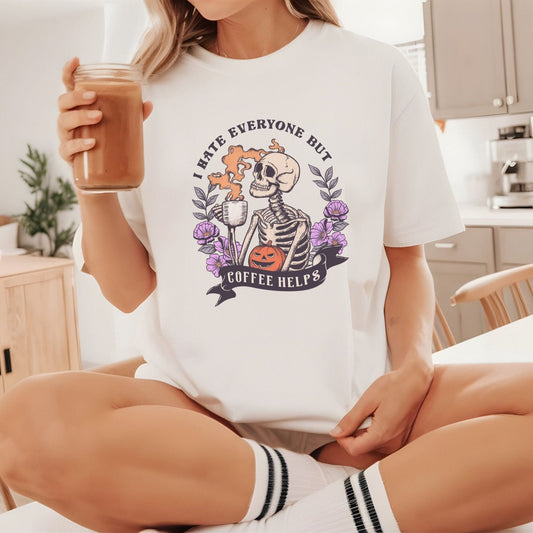“I Hate Everyone But Coffee Helps” t-shirt with skeleton drinking coffee, purple flowers, jack-o’-lantern, and gothic Halloween design.