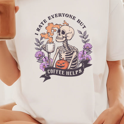 “I Hate Everyone But Coffee Helps” t-shirt with skeleton drinking coffee, purple flowers, jack-o’-lantern, and gothic Halloween design.