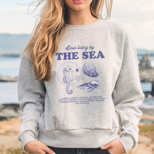 Vintage-style seahorse and seashell graphic with “Slow Living by the Sea” text on a soft unisex sweatshirt, part of the summer coastal collection.